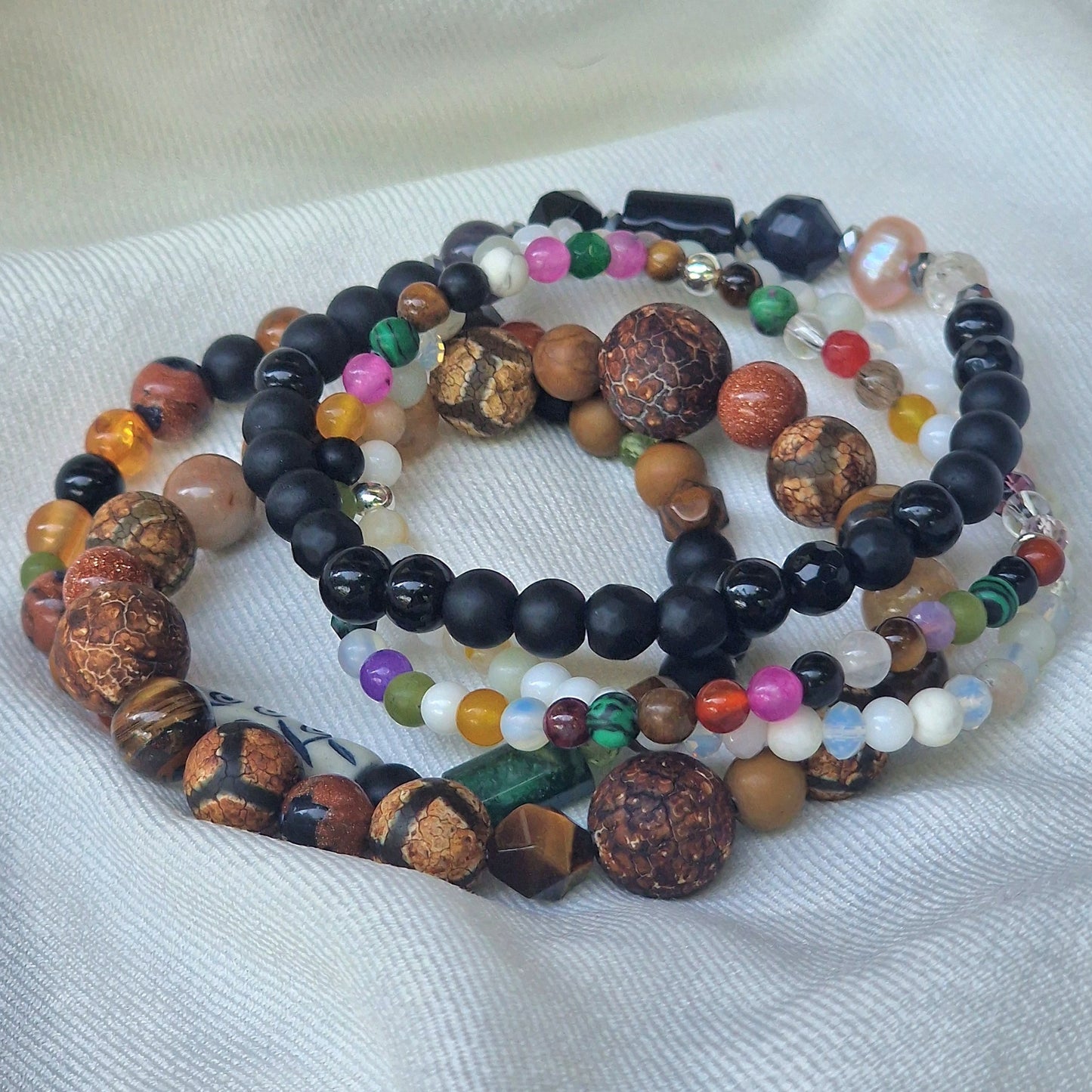 A stack of five Anri Zenya designer gemstone bracelets featuring pearl, agate, howlite, malachite, emerald, topaz, sapphire, tiger’s eye, sunstone and an African Lesotho charm on a neutral background