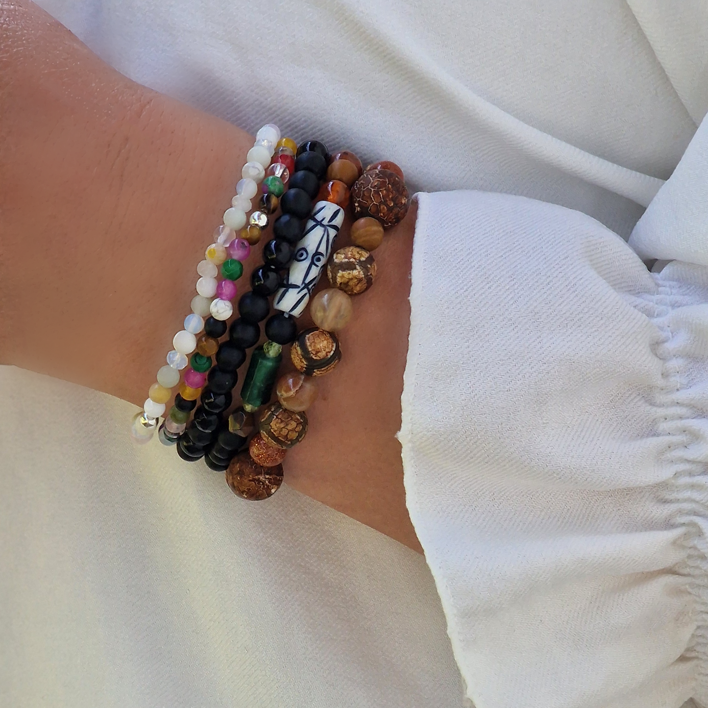 A stack of five Anri Zenya designer gemstone bracelets featuring pearl, agate, howlite, malachite, emerald, topaz, sapphire, tiger’s eye, sunstone and an African Lesotho charm on a neutral background